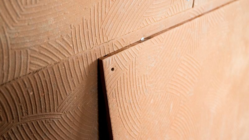 Nahaufnahme einer strukturierten Terrakotta-Fassadenplatte mit eingeprägtem, linearem Muster. Die Platte ist Teil einer montierten Wandverkleidung mit sichtbaren Schraubverbindungen.