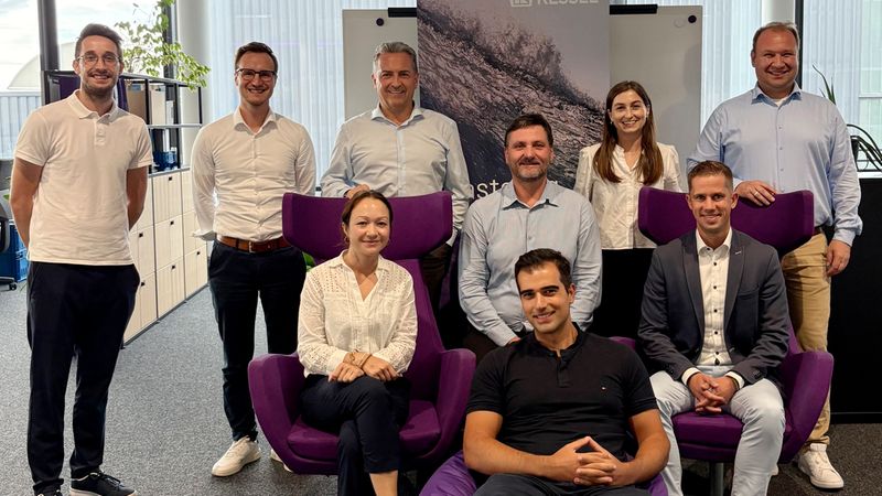 Eine Gruppe von Kessel-Mitarbeitern posiert gemeinsam in einem modernen Büro. Einige Personen stehen, andere sitzen auf modernen, violetten Sesseln und einem Sitzsack, während im Hintergrund ein Firmen-Rollup von Kessel zu sehen ist.