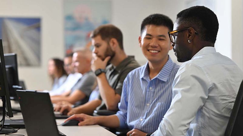 Ein diverses Team von jungen Berufstätigen sitzt in einem modernen Büro an ihren Laptops und arbeitet konzentriert zusammen. Im Vordergrund unterhalten sich zwei Kollegen angeregt, während weitere Mitarbeiter im Hintergrund an ihren Arbeitsplätzen tätig sind.