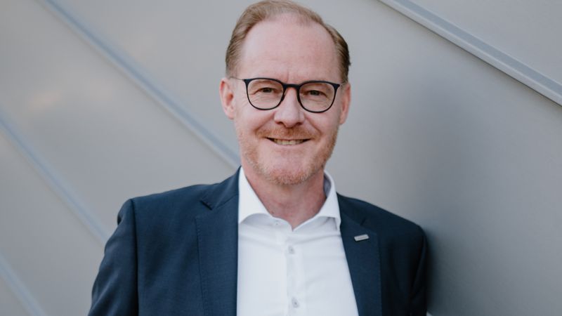 Ein professionelles Business-Porträt eines freundlich lächelnden Mannes mittleren Alters mit Brille, der einen dunkelblauen Anzug und ein weißes Hemd trägt und sich an eine moderne graue Fassade lehnt.