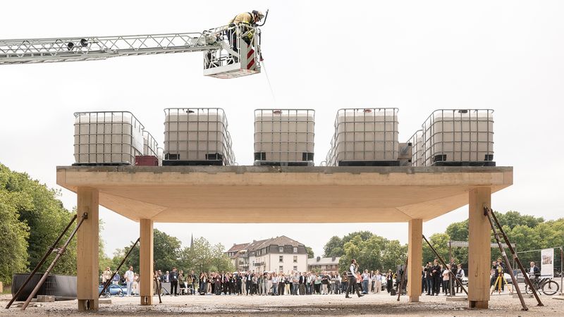 Eine Gruppe von Zuschauern beobachtet eine Vorführung, bei der eine Feuerwehr-Drehleiter Wasser in mehrere IBC-Container füllt, die als Belastungstest auf einem massiven Holzbau-Element platziert wurden.