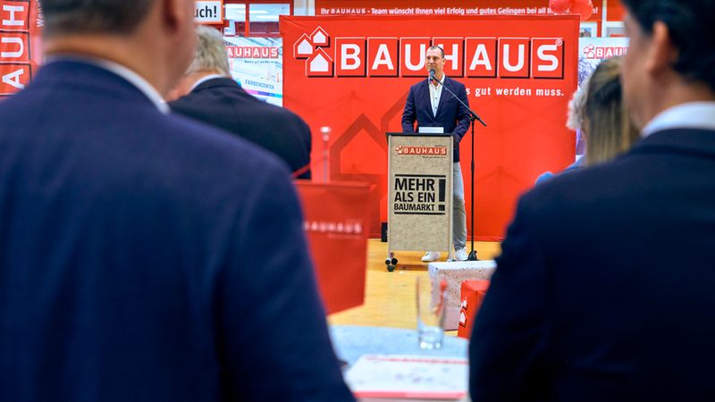 Ein Mann hält eine Präsentation an einem Rednerpult vor einem großen, roten Bauhaus-Banner in einem Baumarkt, während ein Publikum von hinten zuhört.