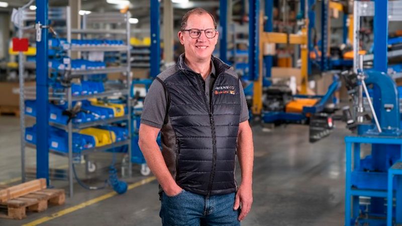 Ein freundlich lächelnder Mitarbeiter steht mit Brille und Weste in einer modernen Produktionshalle vor industriellen Maschinen.