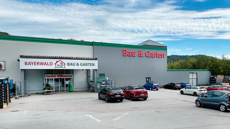 Eine Außenansicht des Bayerwald Bau & Garten Marktes mit einem großen Parkplatz, auf dem mehrere Autos stehen, vor einer Waldkulisse unter blauem Himmel.