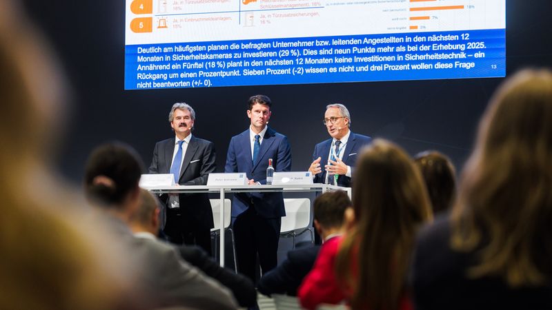Drei Männer im Anzug stehen auf einer Bühne vor einem Publikum und halten eine Präsentation über Statistiken zur Sicherheitstechnik und Investitionsplanung in Unternehmen.