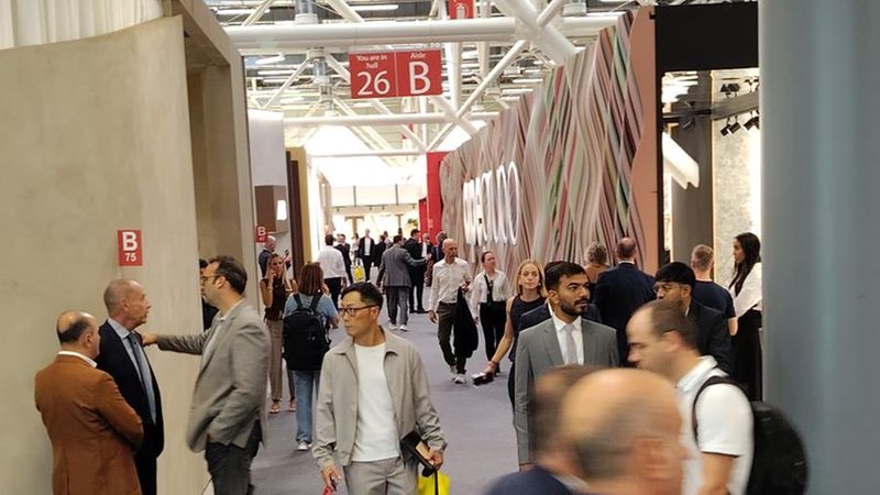 Ein belebter Gang auf einer internationalen Fachmesse, in dem sich zahlreiche Geschäftsleute in förmlicher Kleidung bewegen und unterhalten. Die Architektur des Messegeländes ist durch hohe Decken mit sichtbaren Stahlträgern und Hallenschildern wie '26 B' geprägt, während verschiedene Messestände die Szenerie säumen.