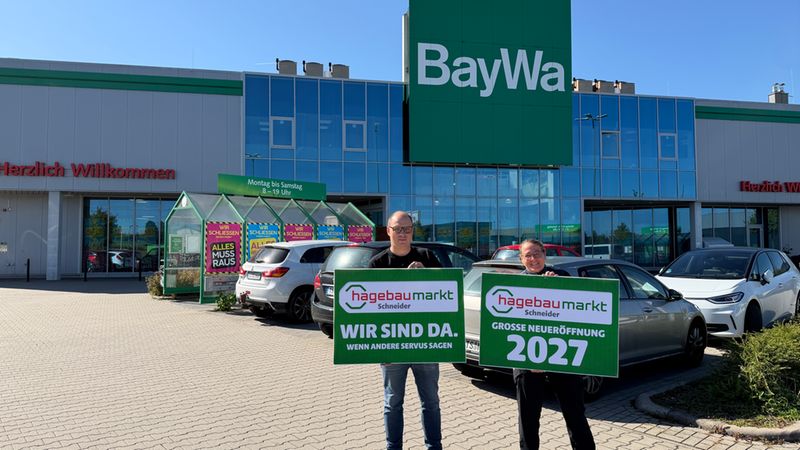 Zwei Personen stehen vor einem BayWa-Gebäude und halten Werbeschilder für die große Neueröffnung eines hagebaumarktes im Jahr 2027.