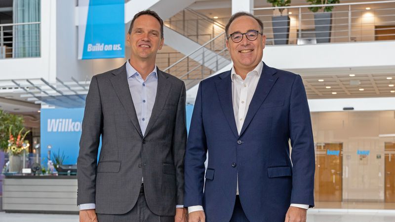 Zwei professionelle Geschäftsmänner im Anzug stehen lächelnd nebeneinander in der hellen, modernen Eingangshalle des Knauf-Unternehmensgebäudes, im Hintergrund sind Firmenschriftzüge wie "Build on us" und "Willkommen" zu sehen.