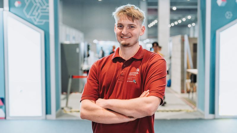 Ein junger Mann mit blonden Haaren trägt ein dunkelrotes Poloshirt mit dem Logo von WorldSkills Germany und steht mit verschränkten Armen lächelnd in einer modernen Messehalle.