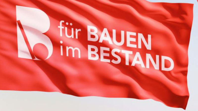 Eine wehende rote Fahne, auf der das Logo und der Schriftzug 'für BAUEN im BESTAND' in weißer Schrift deutlich zu erkennen sind.