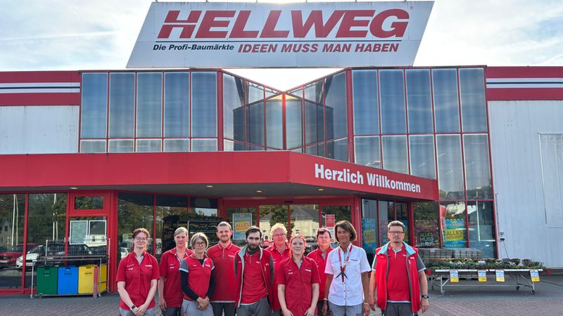 Eine Gruppe von Hellweg Baumarkt Mitarbeitern in einheitlicher Arbeitskleidung posiert gemeinsam vor dem Eingangsbereich einer Hellweg Baumarkt Filiale.