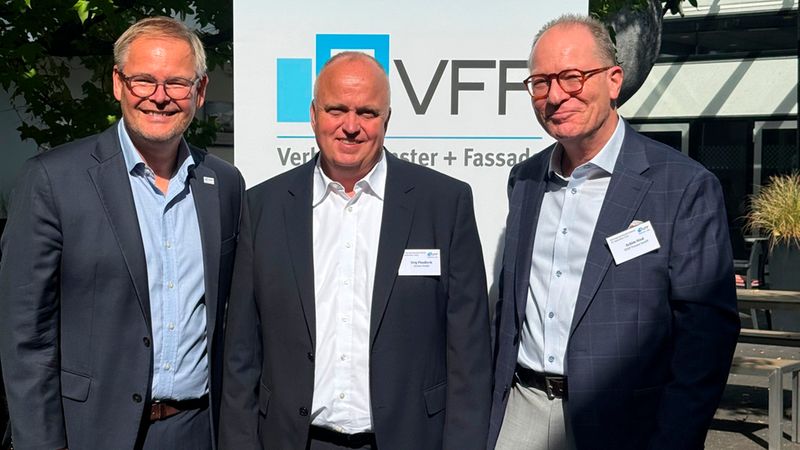 Drei Männer in geschäftlicher Kleidung stehen lächelnd nebeneinander vor einem Banner des Verbandes Fenster + Fassade (VFF) im Freien.