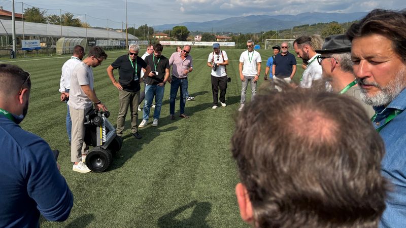 Eine Gruppe von Männern versammelt sich auf einem Fußballplatz, um bei sonnigem Wetter die Vorführung eines automatischen Roboter-Rasenmähers zu beobachten.