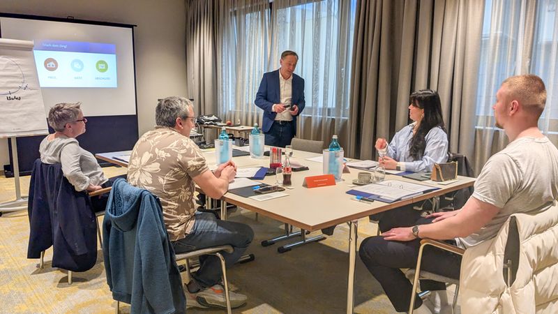 Ein Geschäftstreffen in einem Konferenzraum mit mehreren Personen, die um einen Tisch sitzen und einem Präsentator zuhören.