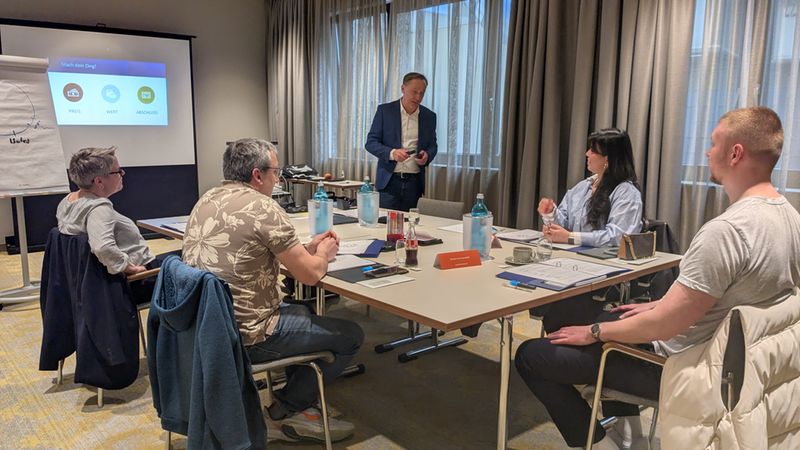 Ein Geschäftstreffen in einem Konferenzraum mit mehreren Personen, die um einen Tisch sitzen und einem Präsentator zuhören.