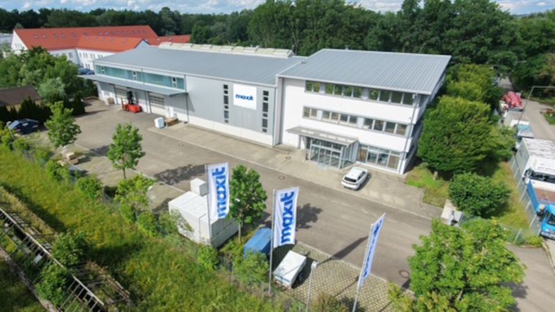 Eine Luftaufnahme des Unternehmensgebäudes der Firma maxit, die den modernen Gebäudekomplex, das Werksgelände mit Firmenfahnen und die umliegende grüne Umgebung zeigt.