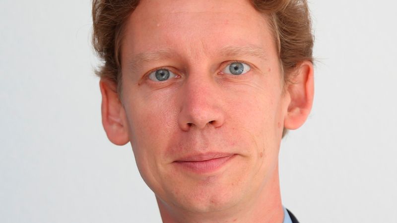 Ein professionelles Portrait eines jungen Mannes mit hellbraunem Haar, der ein hellblaues Hemd und ein dunkelblaues Sakko vor einem neutralen Hintergrund trägt und freundlich in die Kamera blickt.