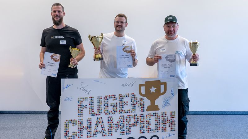 Drei strahlende Gewinner präsentieren stolz ihre Urkunden und Pokale beim Wettbewerb Fugen Champion 2026. Sie halten gemeinsam ein großes Plakat, das mit kleinen Fugenmörtel-Proben künstlerisch gestaltet ist.
