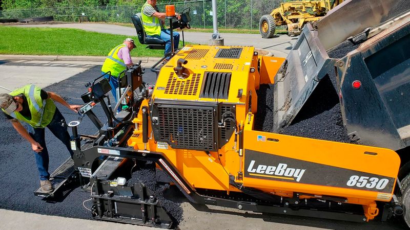 Ein gelber LeeBoy 8530 Strassenfertiger beim Auftragen von frischem Asphalt auf einer Baustelle, unterstützt von Bauarbeitern in Warnwesten.