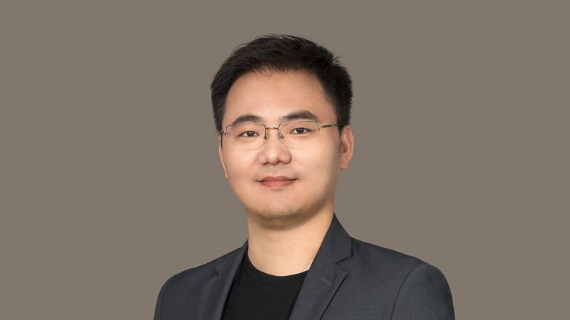 Ein professionelles Porträt eines jungen asiatischen Mannes, der eine Brille, ein schwarzes T-Shirt und ein dunkelgraues Sakko trägt. Er steht vor einem neutralen, grauen Hintergrund und blickt freundlich und selbstbewusst direkt in die Kamera.