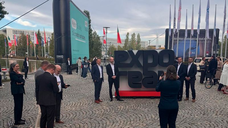 Eine Gruppe von Geschäftsleuten steht vor dem markanten Expo Real Logo auf dem Außenbereich der Immobilienmesse, während eine Frau im Vordergrund das Geschehen fotografiert.