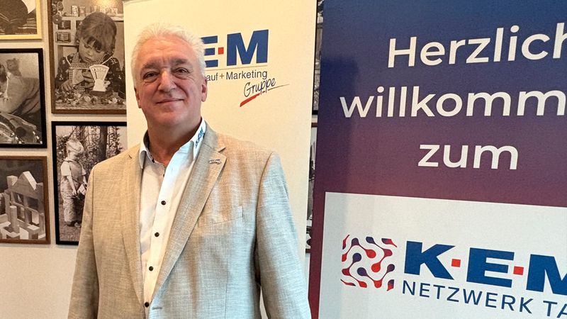 Ein freundlich lächelnder Mann in einem beigen Sakko steht vor einem professionellen Banner der KEM-Netzwerkgruppe. Im Hintergrund sind an der Wand gerahmte Schwarz-Weiß-Fotografien zu sehen.