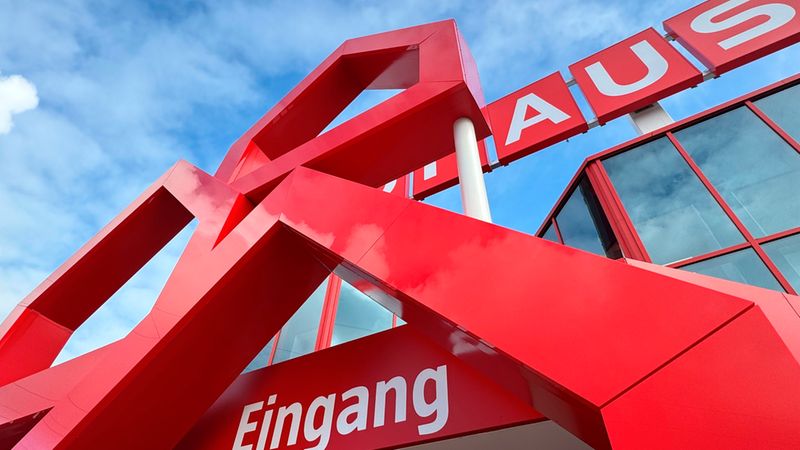 Eine markante Perspektive auf den roten, skulpturalen Eingangsbereich eines Bauhaus-Baumarktes unter einem hellblauen Himmel mit weißen Wolken.