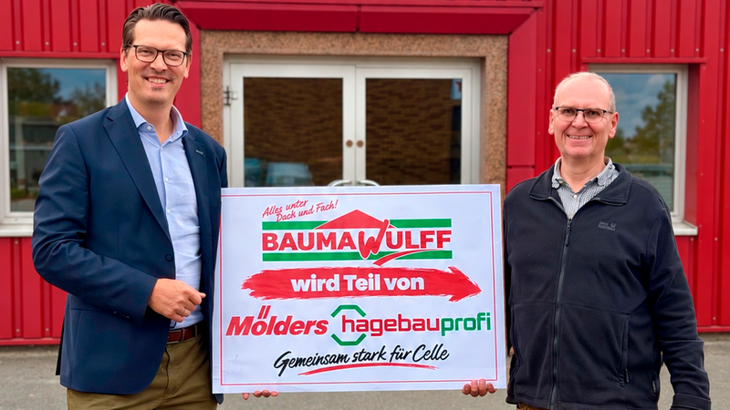 Zwei Männer stehen vor dem roten Gebäude des Baustoffhandels Bauma Wulff und halten gemeinsam ein Schild, das die Übernahme bzw. die Partnerschaft mit Mölders hagebauprofi sowie das Motto "Gemeinsam stark für Celle" präsentiert.