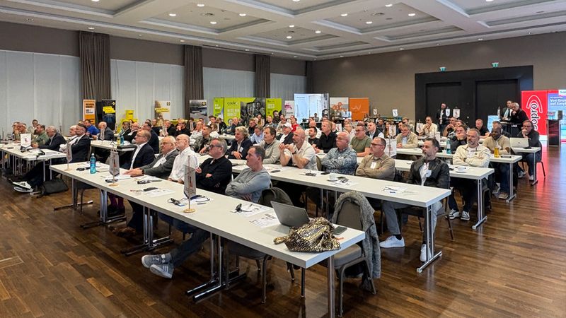 Eine Gruppe von Konferenzteilnehmern sitzt konzentriert in einem modernen, hellen Tagungsraum an langen Tischen und verfolgt eine Präsentation bei einer Veranstaltung.