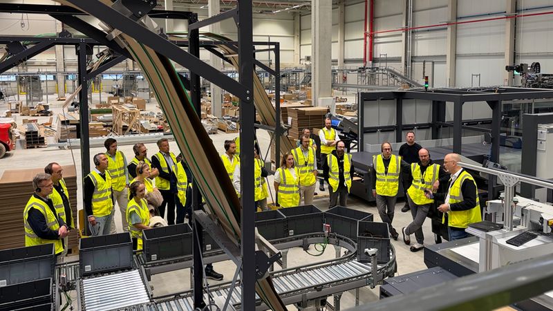 Eine Gruppe von Besuchern trägt neongelbe Warnwesten bei einer professionellen Führung durch eine moderne, hochautomatisierte Logistikhalle mit Fördertechnik und Sortiermaschinen.