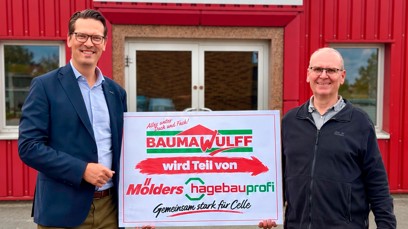 Zwei Männer halten gemeinsam ein Schild vor einem Baustoffhandel-Gebäude, das die Übernahme bzw. Partnerschaft zwischen Bauma Wulff und Mölders hagebauprofi in Celle verkündet.