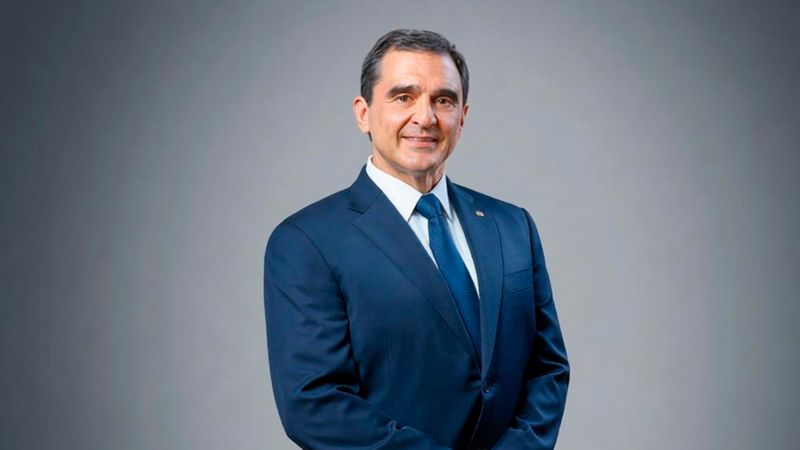 Ein professionelles Porträtfoto von Kostas Skrekas, der einen dunkelblauen Anzug, ein weißes Hemd und eine blaue Krawatte trägt. Er blickt freundlich in die Kamera vor einem neutralen grauen Hintergrund.