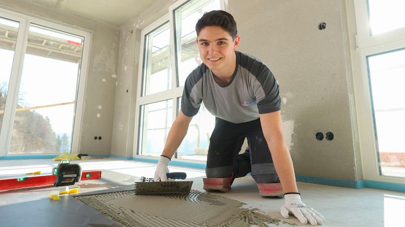Ein freundlich lächelnder junger Fliesenleger kniet auf dem Boden einer hellen Rohbaustelle und trägt mit einer Zahnkelle Fliesenkleber auf den Untergrund auf, während er in die Kamera schaut.