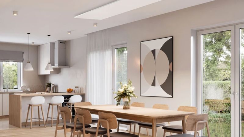 Ein modernes, offenes Wohnkonzept mit einer flachen, weißen Infrarotheizung, die unauffällig an der Decke über einem Essbereich mit Holztisch und Stühlen montiert ist.