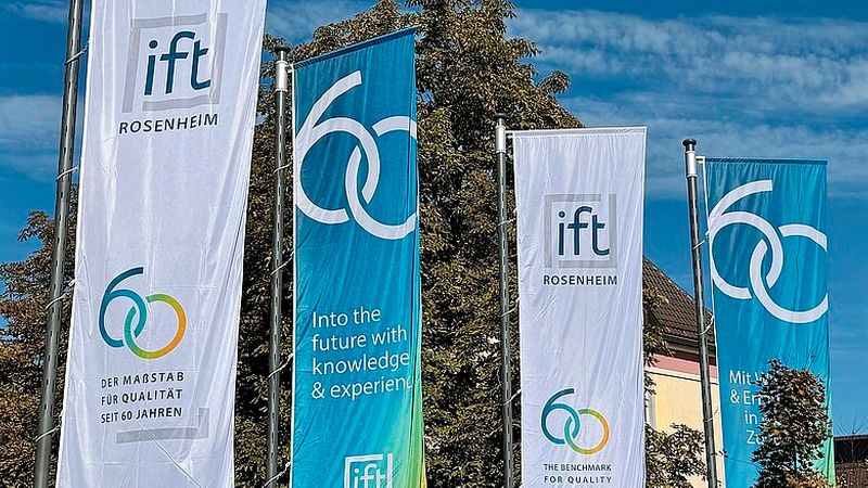 Mehrere Fahnen des ift Rosenheim, die das 60-jährige Jubiläum vor einem Baum und blauem Himmel präsentieren.