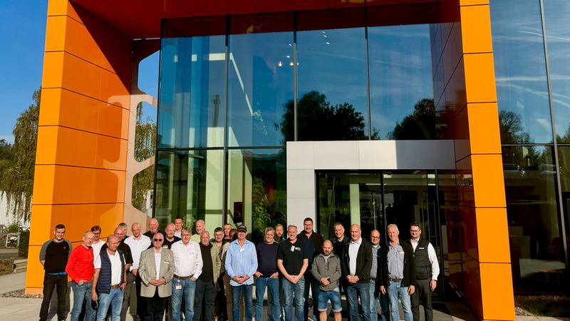 Eine große Gruppe von Mitarbeitern posiert für ein gemeinsames Foto vor einem modernen Bürogebäude mit markanter oranger Fassade und einer großflächigen Glasfront.