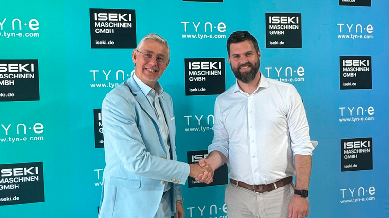 Zwei geschäftlich gekleidete Männer geben sich vor einer türkisblauen Firmenwand mit den Logos von Iseki Maschinen GmbH und Tyn.e einander die Hand, um eine Partnerschaft zu besiegeln.