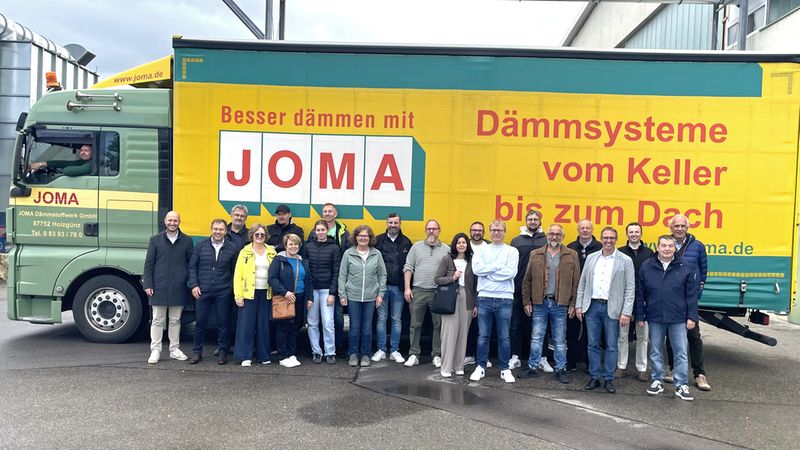 Ein großes Team von Mitarbeitern der JOMA Dämmstoffwerk GmbH posiert gemeinsam vor einem markanten, gelb-grünen Firmen-LKW, der mit dem Slogan „Besser dämmen mit JOMA – Dämmsysteme vom Keller bis zum Dach“ bedruckt ist.