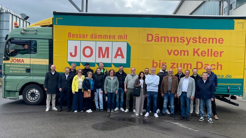 Ein großes Team von Mitarbeitern der JOMA Dämmstoffwerk GmbH posiert gemeinsam vor einem markanten, gelb-grünen Firmen-LKW, der mit dem Slogan „Besser dämmen mit JOMA – Dämmsysteme vom Keller bis zum Dach“ bedruckt ist.