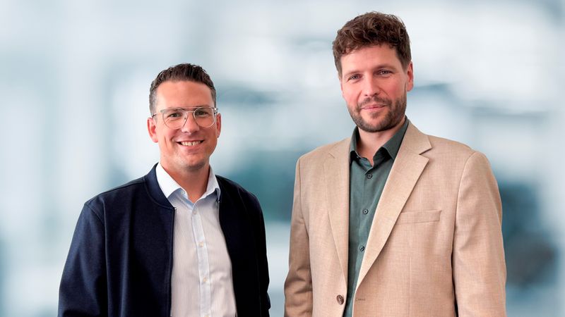 Ein freundliches Porträt von zwei jungen Geschäftspartnern oder Kollegen, die nebeneinander stehen und professionell sowie nahbar in die Kamera lächeln. Beide tragen moderne Business-Casual-Kleidung.