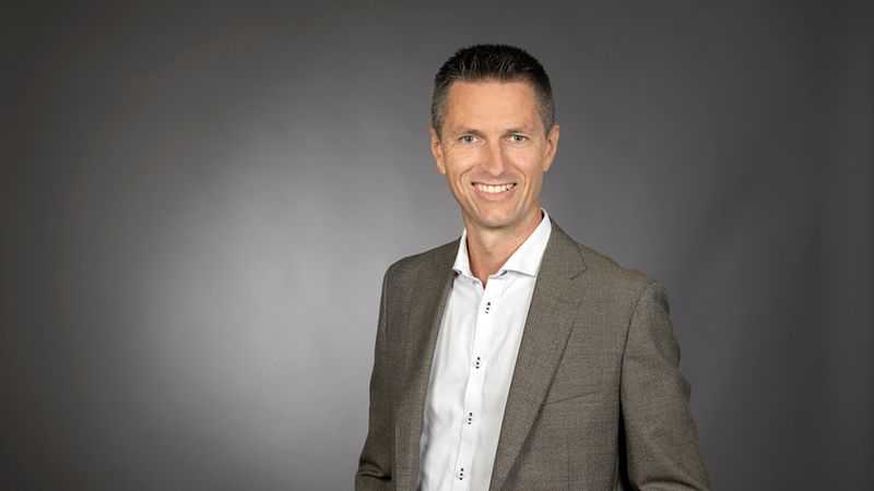 Ein professionelles Portrait eines lächelnden Mannes in einem braunen Sakko und weißem Hemd vor einem neutralen, grauen Hintergrund.