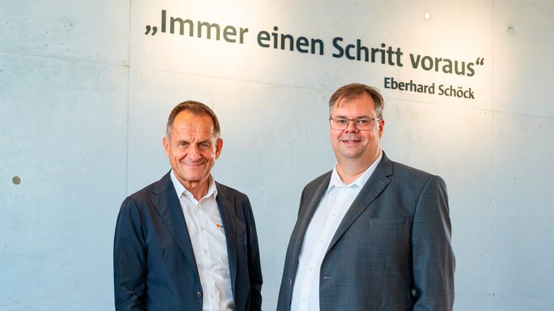 Zwei männliche Führungskräfte in Anzügen stehen selbstbewusst vor einer Betonwand, auf der der Leitsatz „Immer einen Schritt voraus“ von Eberhard Schöck in großen Lettern steht.