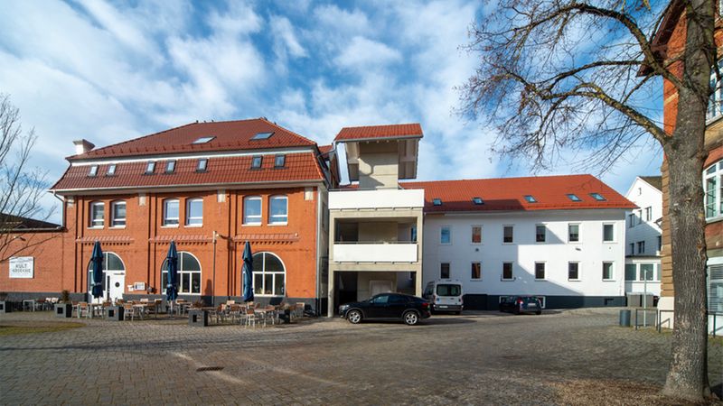 Eine Außenansicht des Restaurants Kult Grieche mit einer Terrasse, die mit Tischen und Stühlen bestuhlt ist, vor einem historischen Backsteingebäude an einem sonnigen Tag.