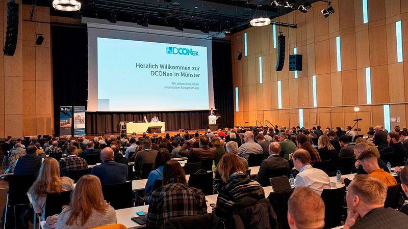 Ein weitläufiger Konferenzsaal mit zahlreichen Teilnehmern, die gespannt auf eine große Leinwand blicken, auf der eine Begrüßungspräsentation für die DCONex in Münster zu sehen ist.