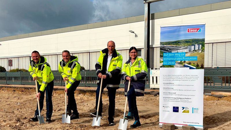 Vier Personen in Warnschutzbekleidung führen einen symbolischen Spatenstich auf einer Baustelle vor einem Tesa-Produktionsgebäude durch. Ein Informationsbanner weist auf das Ziel der klimaneutralen Produktion bis 2030 hin.