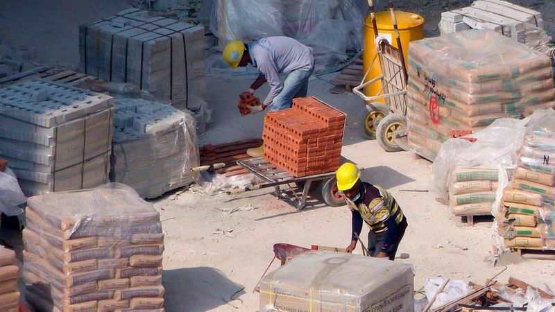 Zwei Bauarbeiter in Schutzkleidung und gelben Helmen arbeiten auf einer aktiven Baustelle, während sie Ziegelsteine auf Paletten und in einer Schubkarre verladen.