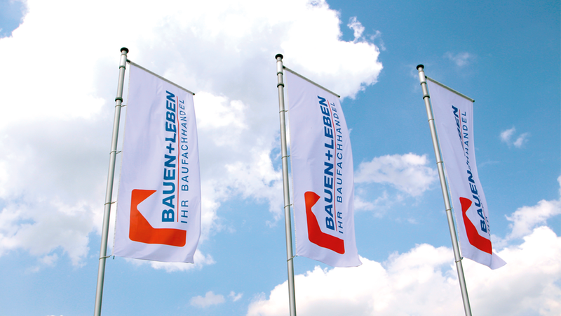 Drei weiße Werbeflaggen mit dem Logo und Schriftzug von BAUEN+LEBEN Ihr Baufachhandel, die an Metallmasten vor einem hellblauen Himmel mit weißen Wolken im Wind wehen.