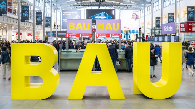 Eine großformatige, gelbe 3D-Skulptur des Schriftzugs BAU dominiert den Vordergrund im hellen Eingangsbereich der Messe München während der Bau-Fachmesse, umgeben von Besuchern und Informationsschaltern.