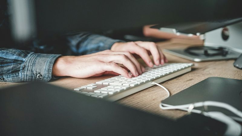 Nahaufnahme von Händen einer Person, die auf einer weißen Computertastatur an einem Holztisch schreibt.