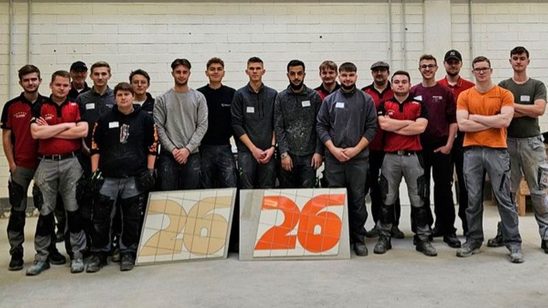 Eine große Gruppe von jungen männlichen Auszubildenden im Handwerk steht in einer Werkstatt. Sie sind in Arbeitskleidung gekleidet und posieren stolz hinter zwei aufgestellten Fliesenarbeiten, auf denen jeweils die Zahl 26 mit Kacheln dargestellt ist.